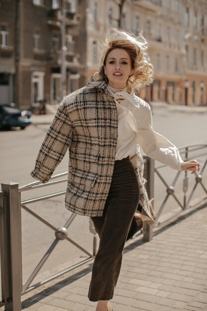 Donna affascinante bionda riccia in eleganti pantaloni marroni, camicetta bianca e cappotto a scacchi che corre nel centro della città city