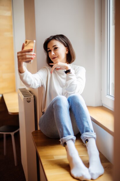 Donna affascinante che si siede sul davanzale in blue jeans con il telefono che fa selfie