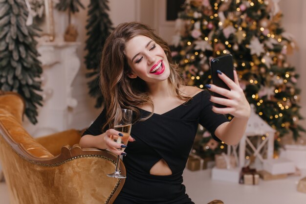 Donna affascinante con manicure alla moda che fa selfie con champagne vicino all'albero di Natale. Ritratto dell'interno del modello femminile bianco sveglio in abbigliamento del partito che cattura maschera di se stessa in vacanza.