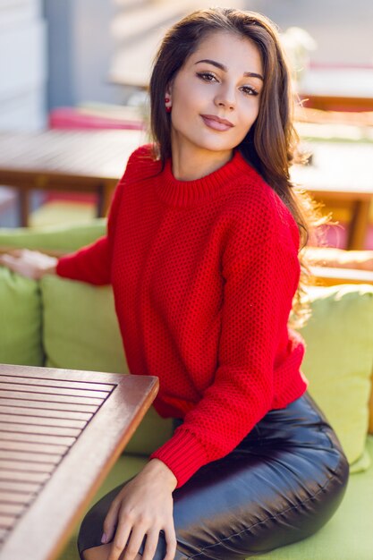 Donna affascinante del brunette in maglione lavorato a maglia autunno rosso e gonna di cuoio che si distendono sul sofà nel ristorante dello spazio aperto.