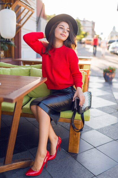 Donna affascinante del brunette in maglione lavorato a maglia autunno rosso e gonna di cuoio che si distendono sul sofà nel ristorante dello spazio aperto.
