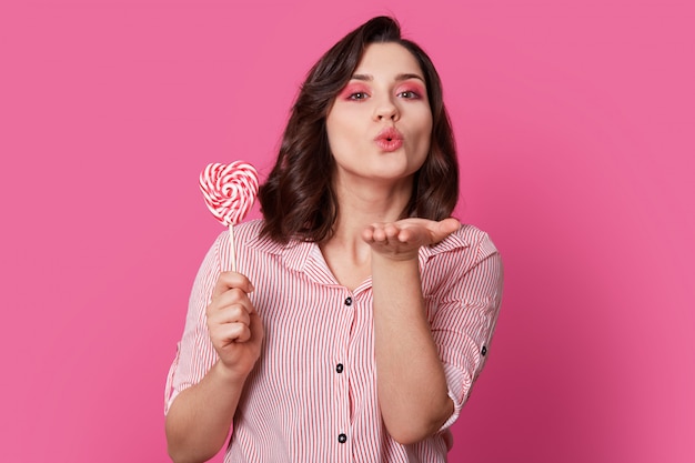 donna affettuosa soffia bacio d'aria, vestita con abiti alla moda, tiene caramelle, ha un trucco luminoso, posa su roseo. Il suo amore è per te. La bella ragazza pinup si diverte al coperto.