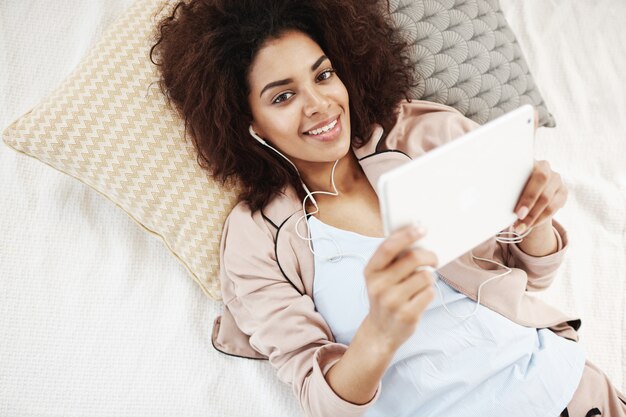 Donna africana allegra in indumenti da notte e compressa sorridente della tenuta della cuffia che si trova sul letto.