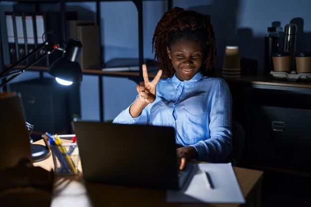 Donna africana che lavora in ufficio di notte sorridendo guardando la telecamera mostrando le dita facendo il segno di vittoria numero due