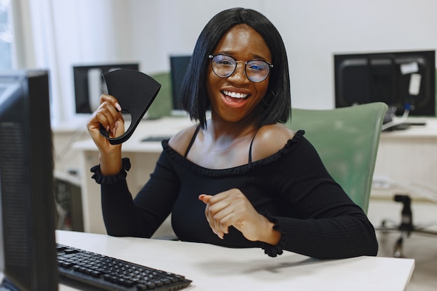 Donna africana che si siede nella classe di informatica. La signora con gli occhiali sorride alla telecamera. Studentessa seduta al computer.