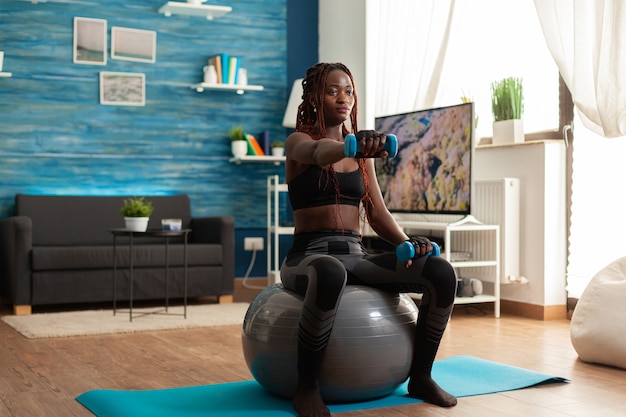 Donna africana che usa la palla di stabilità tenendo le braccia tese allenando le spalle usando manubri blu, nel soggiorno di casa per modellare i muscoli e uno stile di vita sano, vestita con abbigliamento sportivo