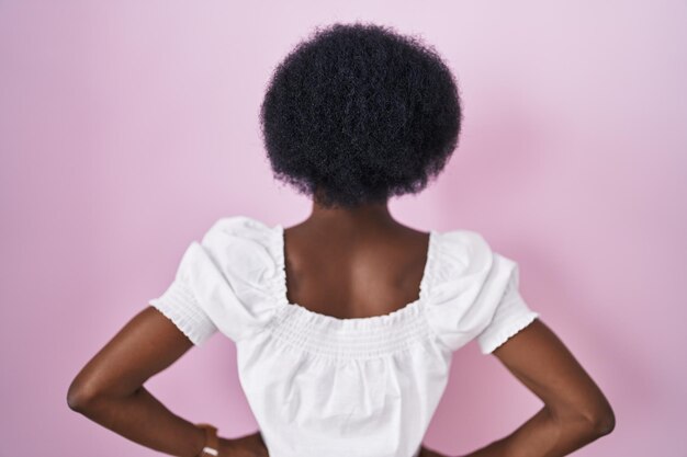 Donna africana con capelli ricci in piedi su sfondo rosa in piedi all'indietro guardando lontano con le braccia sul corpo