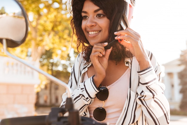 donna africana felice che si siede sulla motocicletta moderna e fissa il suo moto halmet all'aperto