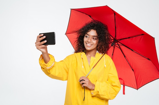 Donna africana felice in impermeabile che si nasconde sotto l'ombrello e che fa selfie sullo smartphone sopra fondo bianco