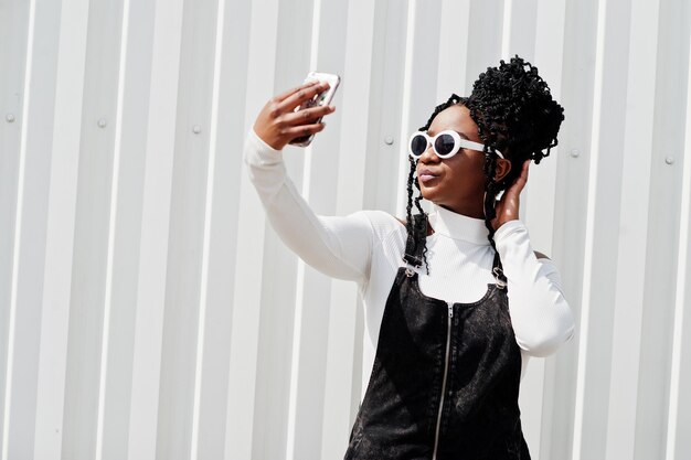 Donna africana in tuta di jeans gonna occhiali da sole bianchi posati contro il muro di acciaio bianco e facendo selfie per telefono