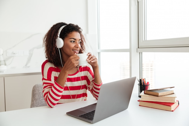 Donna africana sorridente che si siede dalla tavola con il computer portatile