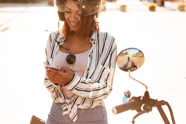 Donna africana sorridente che si siede sulla motocicletta moderna all'aperto e che per mezzo del suo smartphone