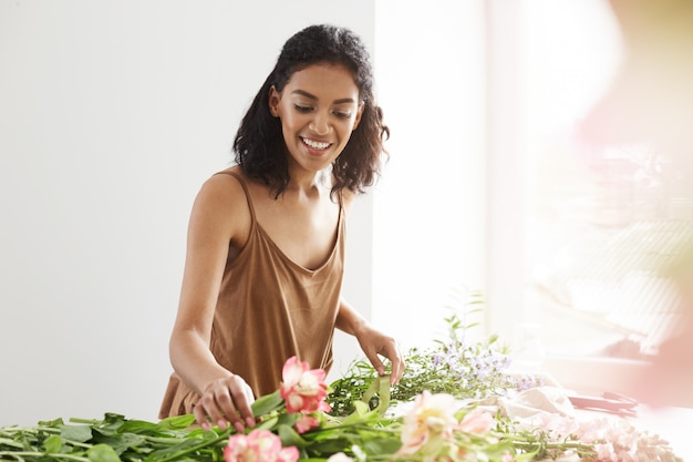 Donna africana tenera felice che sorride facendo mazzo dei fiori nel luogo di lavoro sopra la parete bianca.