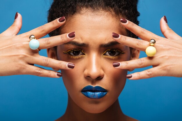 Donna afro concentrata alla moda con trucco variopinto che dimostra gli anelli sulle sue dita che tengono le mani al fronte, isolato sopra la parete blu