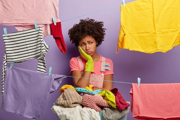 Donna afro stanca sconvolta occupata con i lavori domestici, indossa guanti di gomma, asciuga i vestiti, ha molti lavori in casa, si trova vicino al cesto di biancheria sporca, isolato su sfondo viola.