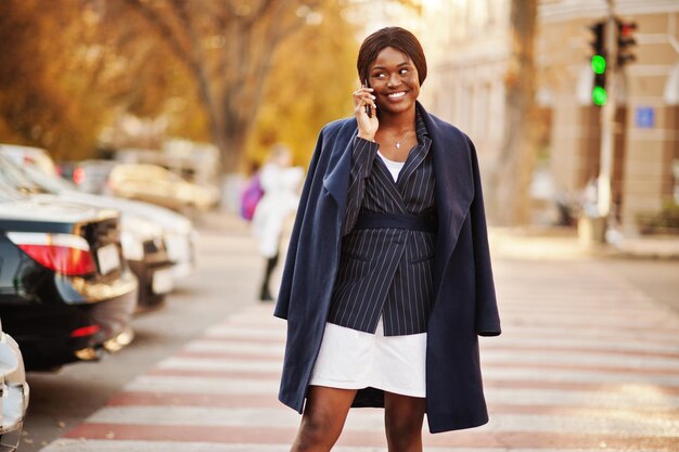 Donna afroamericana alla moda di successo in cappotto che cammina al passaggio pedonale e parla al telefono