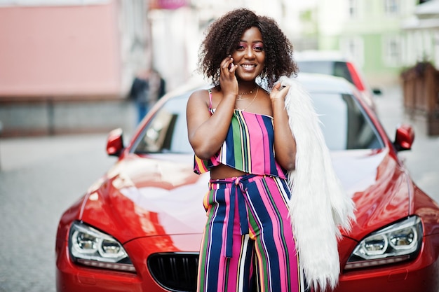 Donna afroamericana alla moda in tuta a righe rosa con soffice pelliccia sintetica posata contro un'auto di lusso rossa ricca e parlando al telefono cellulare