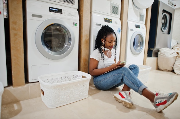 Donna afroamericana allegra che si siede con gli auricolari e legge la rivista vicino alla lavatrice nella lavanderia self-service