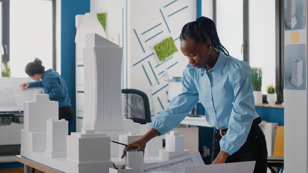 Donna afroamericana che analizza il piano dei progetti e il modello di edificio per lavorare sull'architettura. Architetto che lavora con maquette e stampa di layout per progettare la costruzione urbana per lo sviluppo.