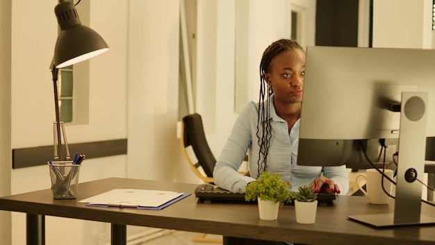 Donna afroamericana che fa lavoro esecutivo sul computer durante l'ora del tramonto. Lavorare su presentazioni aziendali per inviare report di dati via e-mail, utilizzando ricerche web online e documenti cartacei.