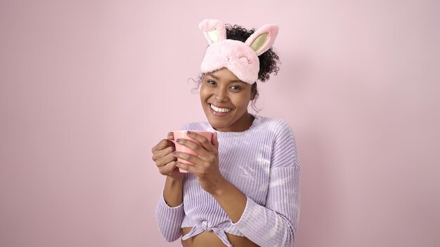 Donna afroamericana che indossa la maschera del sonno che beve una tazza di caffè su sfondo rosa isolato