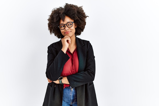 Donna afroamericana con capelli afro che indossa giacca da lavoro e occhiali con la mano sul mento che pensa alla domanda, espressione pensierosa. volto sorridente e pensieroso. concetto di dubbio.