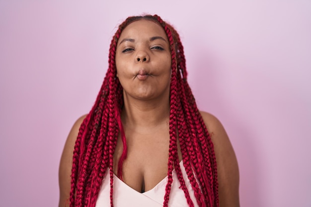 Donna afroamericana con capelli intrecciati in piedi su sfondo rosa che fa la faccia di pesce con le labbra, gesto folle e comico. espressione buffa.