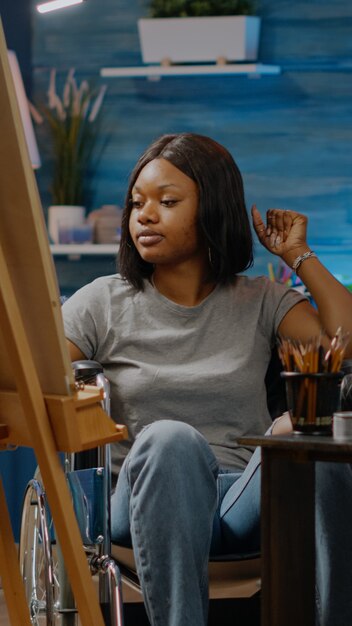 Donna afroamericana con handicap che lavora su un progetto artistico mentre è seduta nello spazio studio. Artista nero invalido con disabilità in sedia a rotelle che disegna un vaso bianco su tela e cavalletto