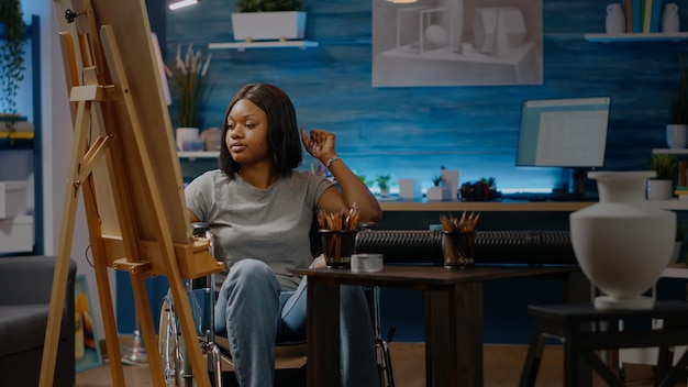 Donna afroamericana con handicap che lavora su un progetto artistico mentre è seduta nello spazio studio. Artista nero invalido con disabilità in sedia a rotelle che disegna vaso bianco su tela e cavalletto