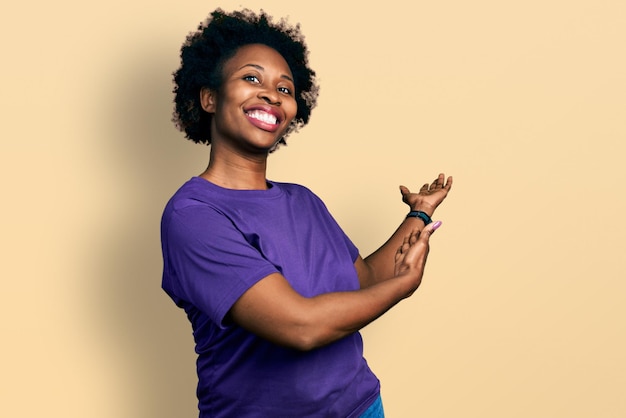 Donna afroamericana con i capelli afro che indossa una maglietta viola casual che invita a entrare sorridendo naturale con la mano aperta