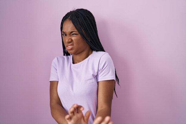 Donna afroamericana con trecce in piedi su sfondo rosa espressione disgustata, scontenta e timorosa che fa il viso disgustato perché reazione di avversione.
