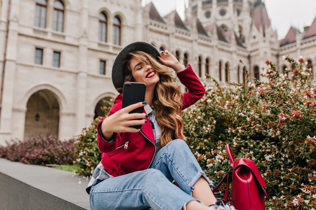 Donna agghiacciante in jeans retrò facendo selfie con gli occhi chiusi vicino a fiori di strada