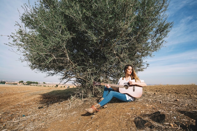 Donna alla moda che gioca chitarra vicino al cespuglio