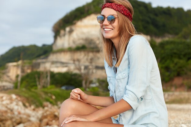 Donna alla moda con gli occhiali da sole che si siedono sulla spiaggia