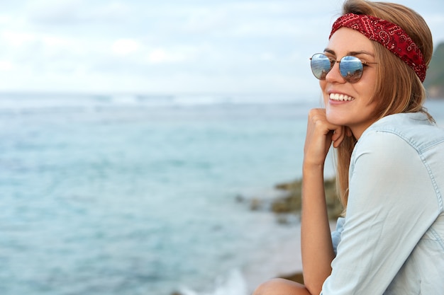 Donna alla moda con gli occhiali da sole che si siedono sulla spiaggia