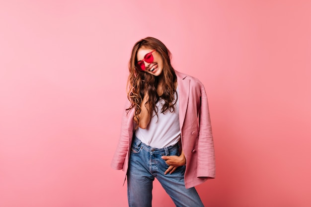 Donna alla moda con risata felice dei capelli lunghi dello zenzero. Ritratto dell'interno di beata ragazza caucasica in occhiali da sole alla moda e giacca rosa.