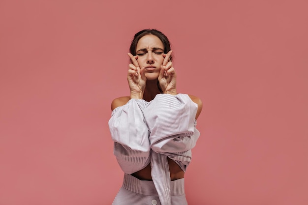 Donna alla moda dai capelli neri in camicetta a righe chiare e gonna fresca incrociando le dita e posando con gli occhi chiusi su sfondo rosa