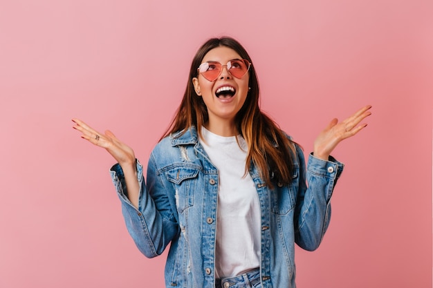 Donna alla moda emotiva che osserva in su su sfondo rosa. Ragazza bruna sorpresa in giacca di jeans in posa con la bocca aperta.