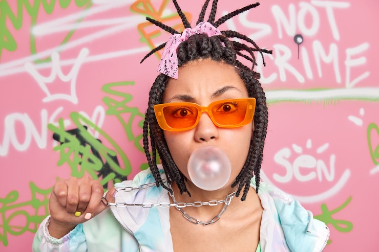 Foto gratuito donna alla moda fresca con trecce di capelli soffia gomme da masticare tiene catena di metallo appesa al collo vestita con abiti alla moda pone un muro di graffiti colorati