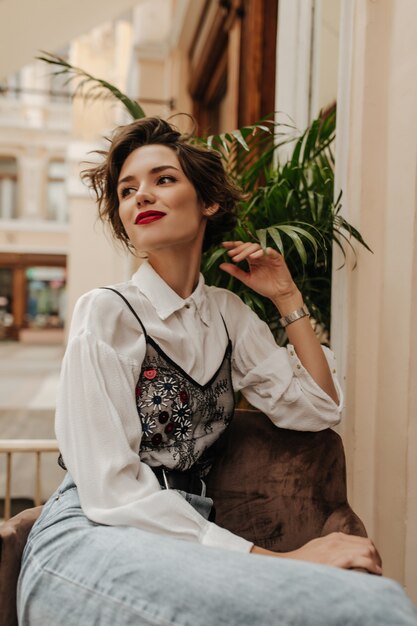 Donna alla moda in camicetta bianca e jeans che sorridono nella caffetteria. Donna alla moda con i capelli corti del brunette che si siede nel ristorante.