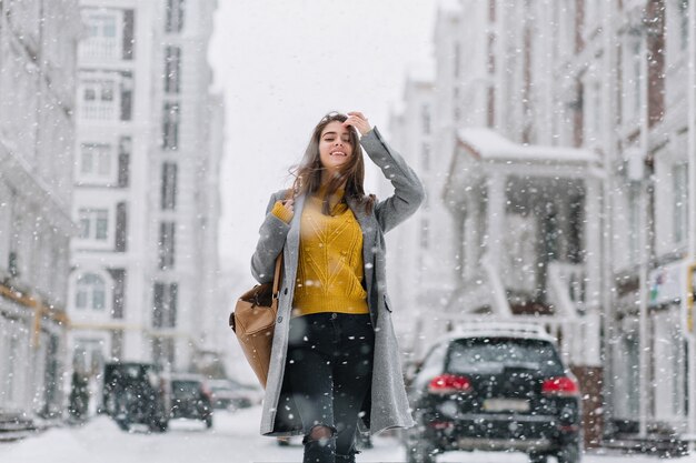 Donna alla moda in maglione giallo lavorato a maglia in posa sotto la nevicata sulla strada. Ritratto all'aperto di adorabile signora in cappotto grigio che gode della neve