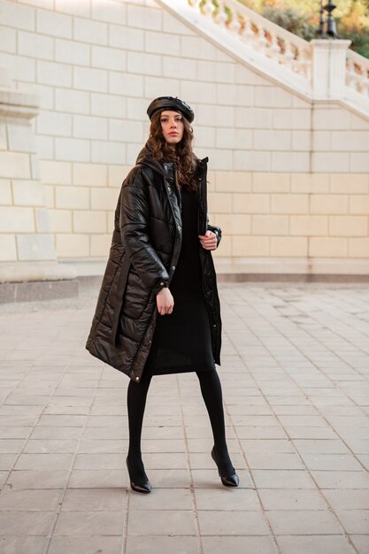 Donna alla moda in posa in inverno autunno moda tendenza nero piumino e berretto in pelle berretto in vecchia bella strada indossando scarpe tacco alto