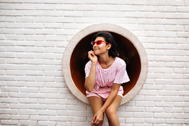 Donna alla moda in posa sul muro di mattoni. Colpo esterno di donna allegra in occhiali da sole rosa.