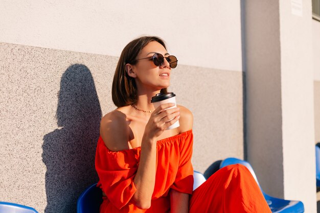 Donna alla moda in vestiti arancioni al tramonto allo stadio della pista ciclabile con una tazza di caffè