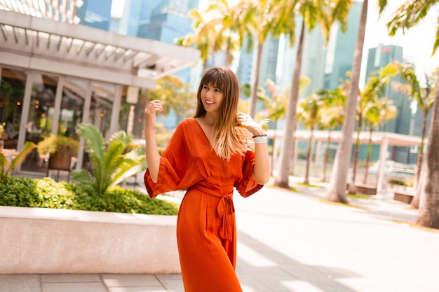 Donna alla moda in vestito arancio che posa sulla passeggiata con le palme e i grattacieli in grande città moderna