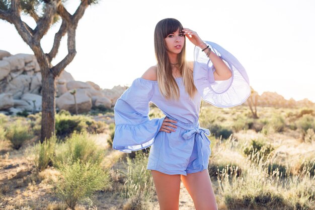 Donna alla moda in vestito blu elegante con maniche larghe in posa nel parco nazionale di Joshua tree. Incredibile tramonto morbido.