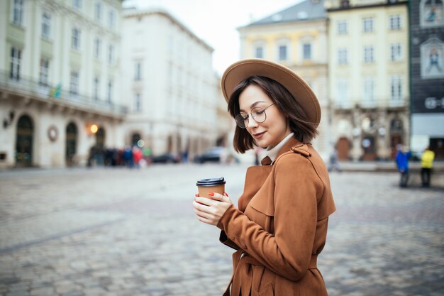 Donna alla moda in vetri che beve caffè sul fondo della città di autunno