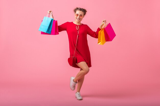 Donna alla moda sorridente felice attraente shopaholic in vestito alla moda rosso che tiene le borse della spesa colorate sul muro rosa isolato, vendita eccitata, tendenza moda primavera estate