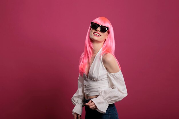 Donna alla moda spensierata che indossa occhiali da sole alla moda sulla fotocamera, sentendosi sicura e attraente su sfondo rosa. Bella ragazza sensuale con occhiali carini sul viso, acconciatura funky.