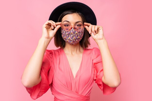 Donna alla moda vestita maschera protettiva elegante. Indossare cappello nero e occhiali da sole. In posa sul muro rosa